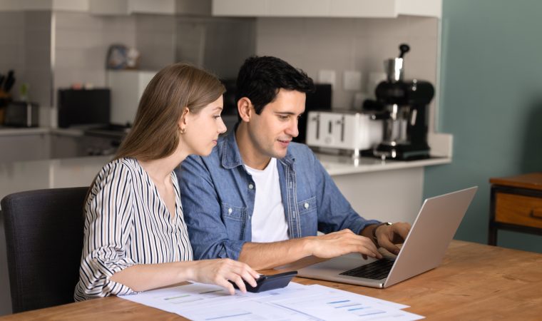 Focused young married couple managing family budget, check finances seated at table in modern kitchen, paying bills through secure e-banking system using laptop, make payment and virtual transactions