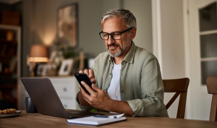 A man working from home over a laptop and using a mobile phone sitting at the wooden table.